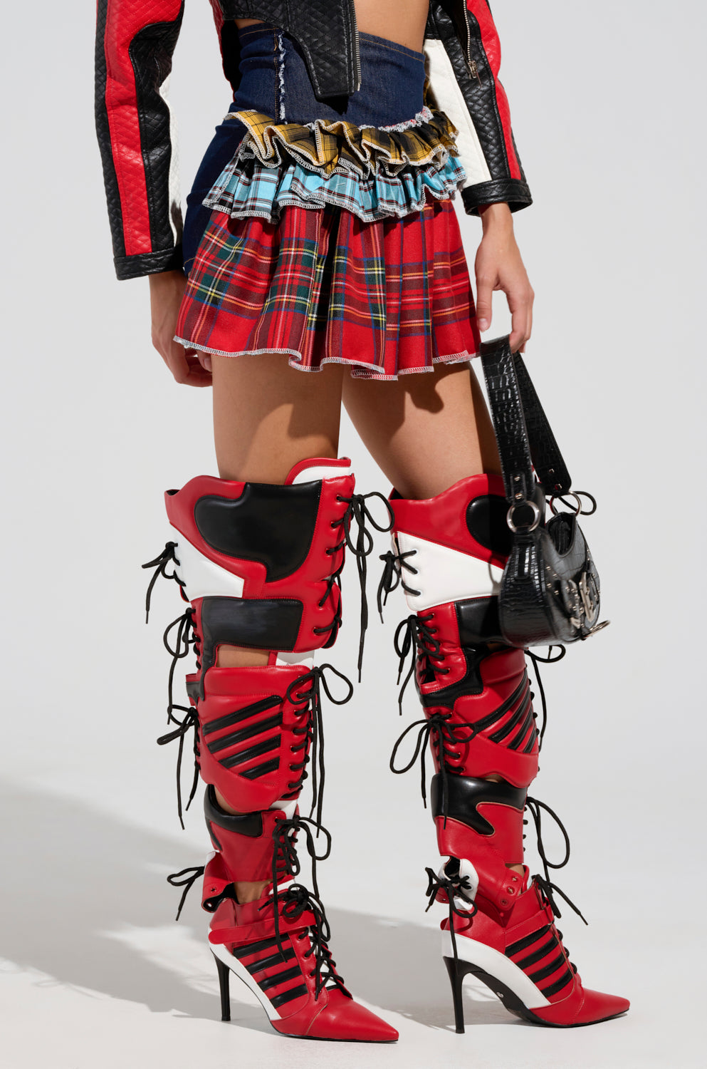 A person showcases the Call Me Back Denim Skirt with Plaid Detail, highlighted by a red and black design. They pair it with eye-catching red over-the-knee boots adorned with black lace-ups and a sophisticated black purse. The jacket complements the look with its bold black and red panels.