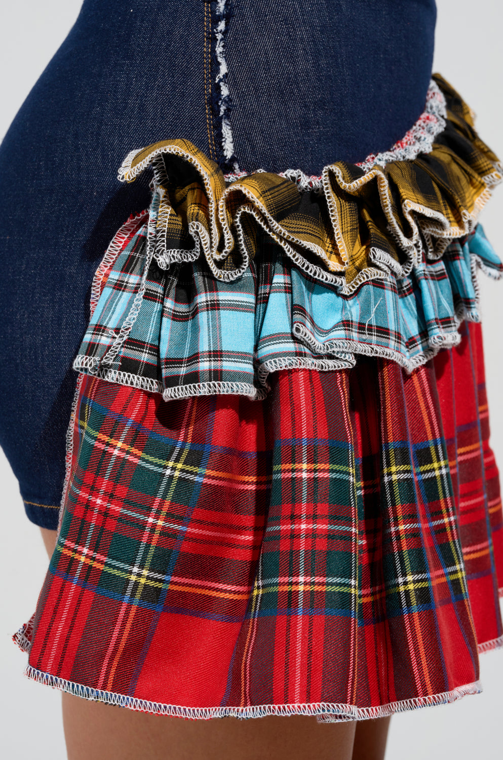 A close-up of the CALL ME BACK DENIM SKIRT WITH PLAID DETAIL showcases its ruffled layers in diverse plaid designs, including shades of red, blue, and yellow. The skirt's asymmetrical shape and textured fabric create a lively contrast with the denim.