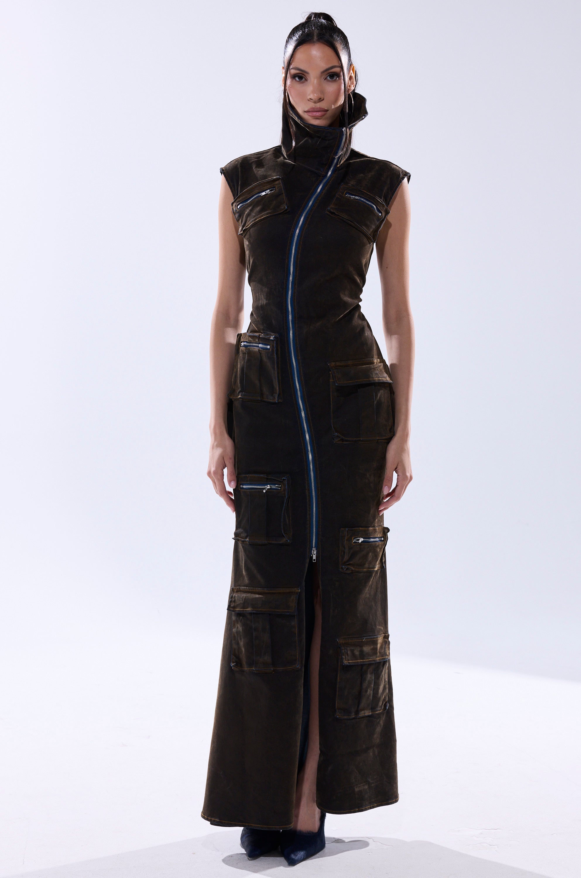 A woman wears the NOW YOU SEE ME VELVET DENIM MAXI DRESS—a long, sleeveless dark brown dress with cargo pockets and a striking blue diagonal zipper—standing against a plain light background.