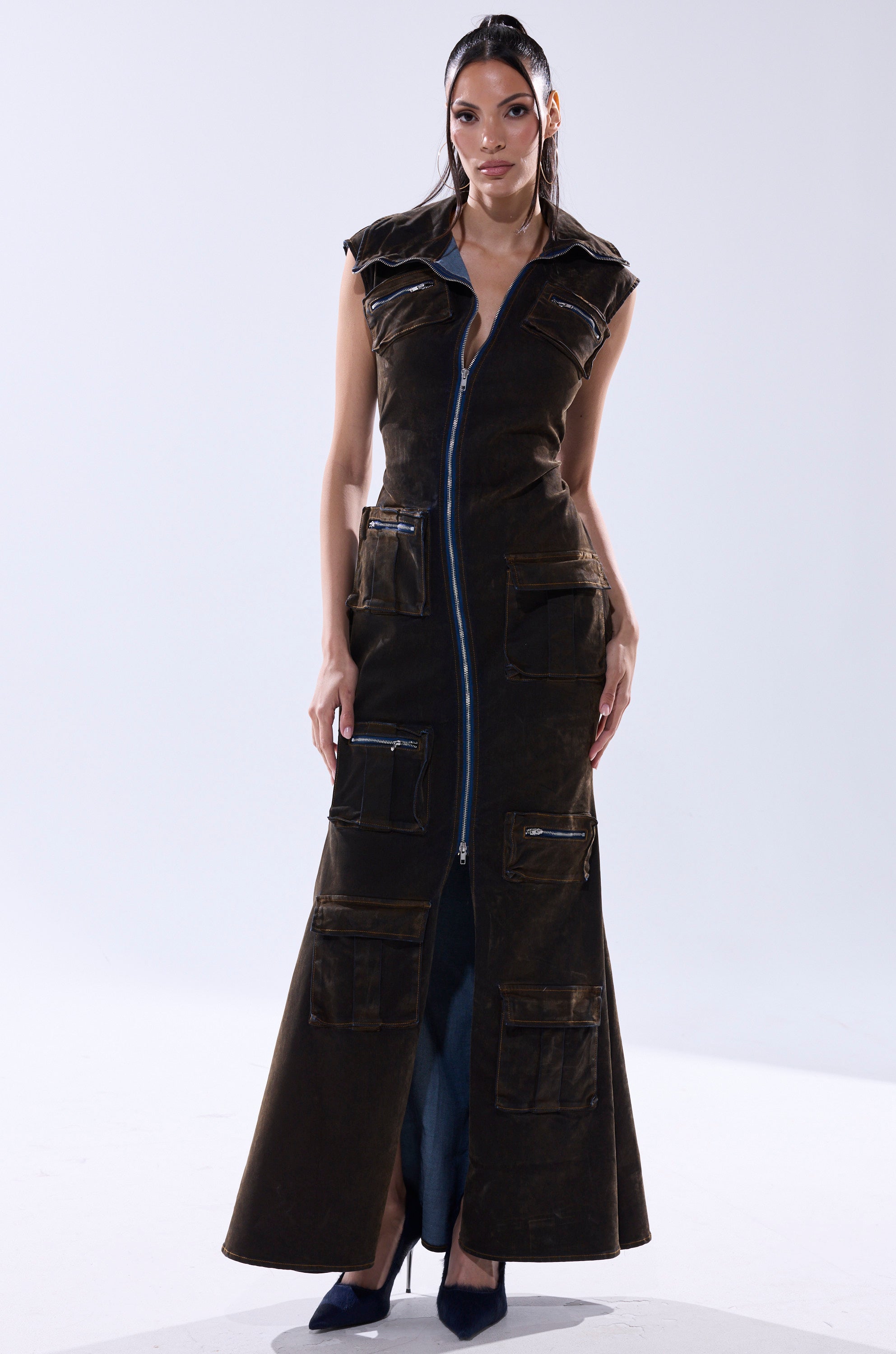 A woman faces forward in the NOW YOU SEE ME VELVET DENIM MAXI DRESS—a long, dark brown velvet denim piece with a blue front zipper and multiple cargo pockets. She wears hoop earrings, black pointed heels, and has straight dark hair against a white background.