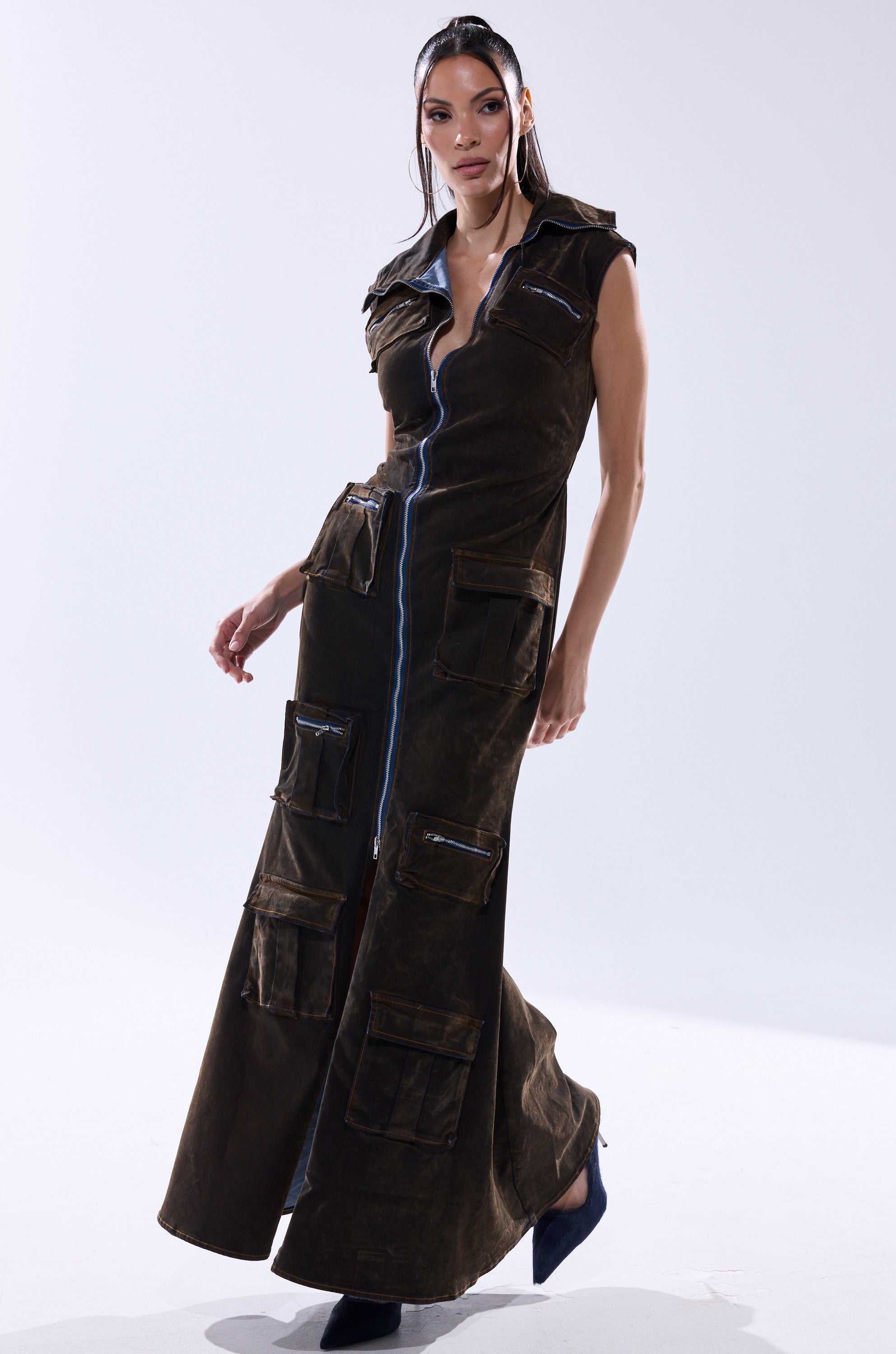 A woman models the NOW YOU SEE ME VELVET DENIM MAXI DRESS—sleeveless, floor-length, with cargo pockets and a central blue zipper—paired with dark heels and styled straight-back hair against a plain white background.