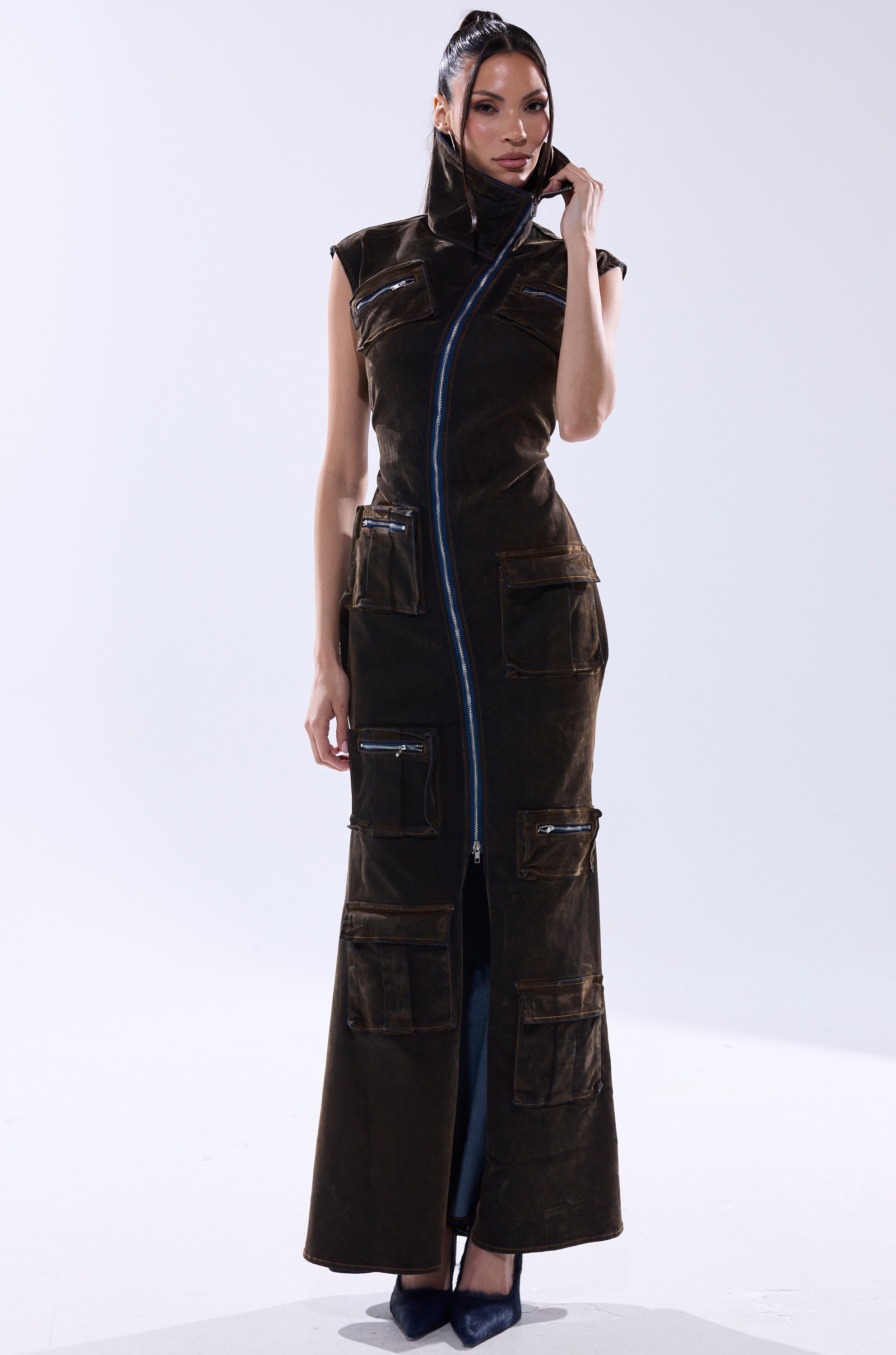 A woman models the NOW YOU SEE ME VELVET DENIM MAXI DRESS, a long dark brown sleeveless dress with cargo pockets and a high zip-up collar, posing against a plain light background.