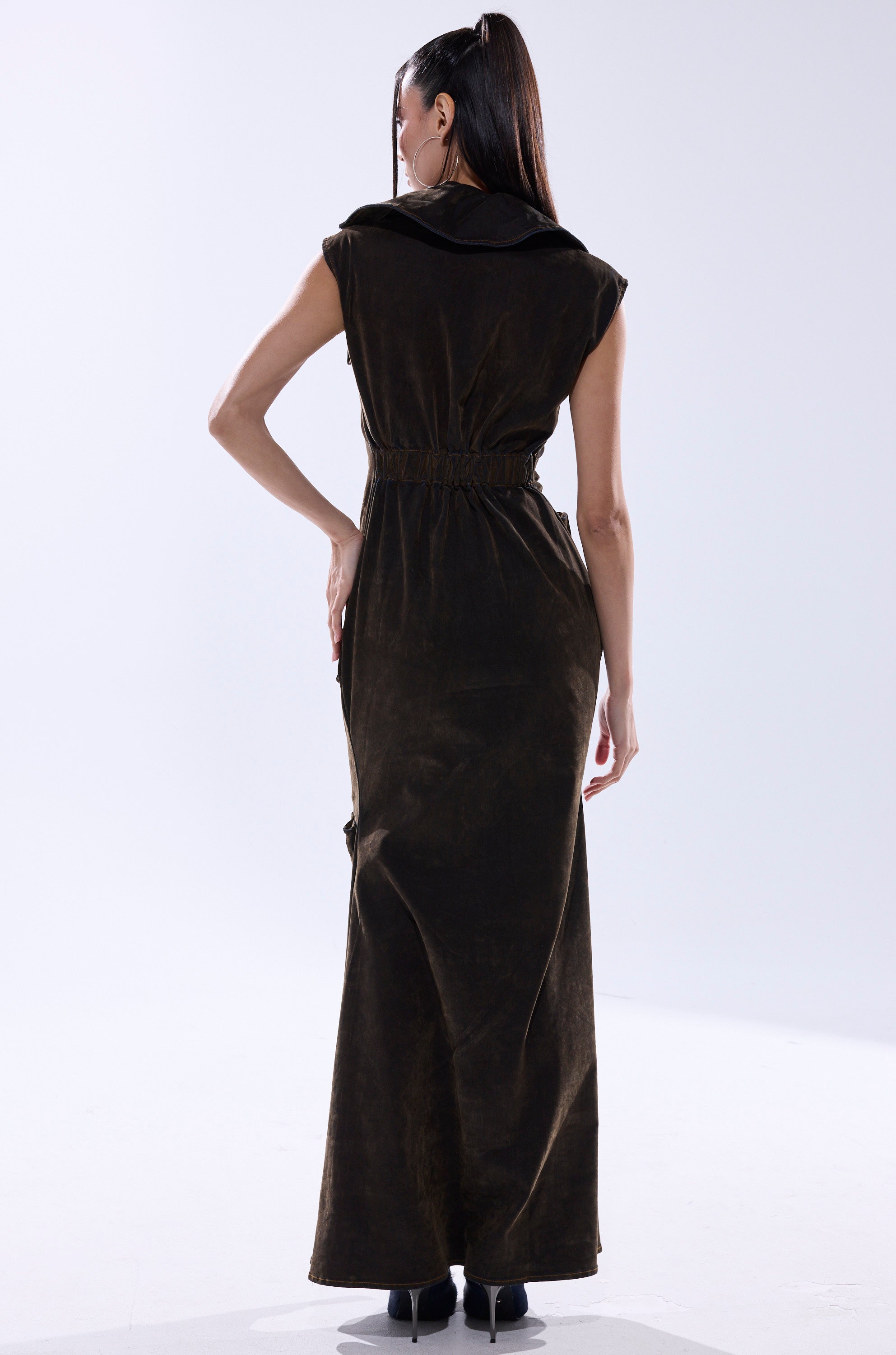 A woman faces away in the NOW YOU SEE ME VELVET DENIM MAXI DRESS, featuring a sleeveless, dark brown velvet look with a loose collar and fitted waist, paired with black heels against a plain light background.