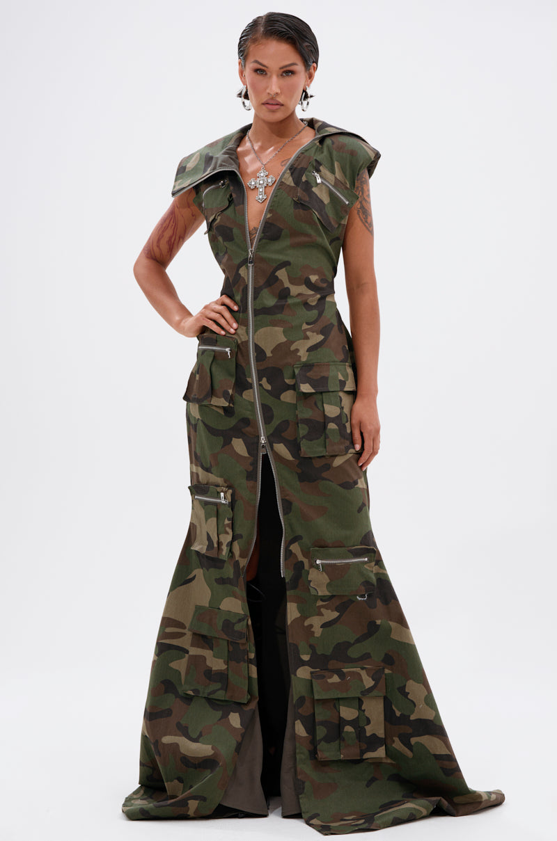 A person stands facing forward wearing the NOW YOU SEE ME CAMO PRINT MAXI DRESS, featuring a hood, large front zipper, and multiple cargo pockets. They accessorize with tattoos, earrings, and a statement necklace.
