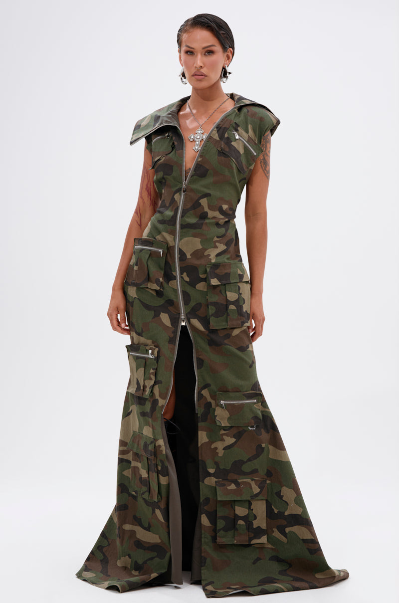 A woman wears the NOW YOU SEE ME CAMO PRINT MAXI DRESS, a sleeveless maxi with front zipper, cargo pockets, and flared hem. She sports short wet-look hair, visible tattoos, and a statement necklace.