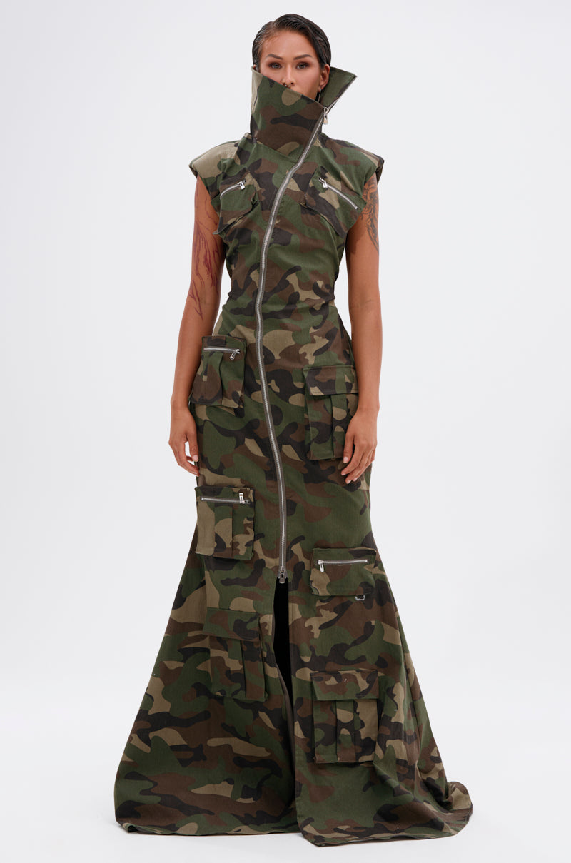 Wearing the NOW YOU SEE ME CAMO PRINT MAXI DRESS, featuring a high zipped collar, cargo pockets, and flared hem, a person stands against a plain white background.