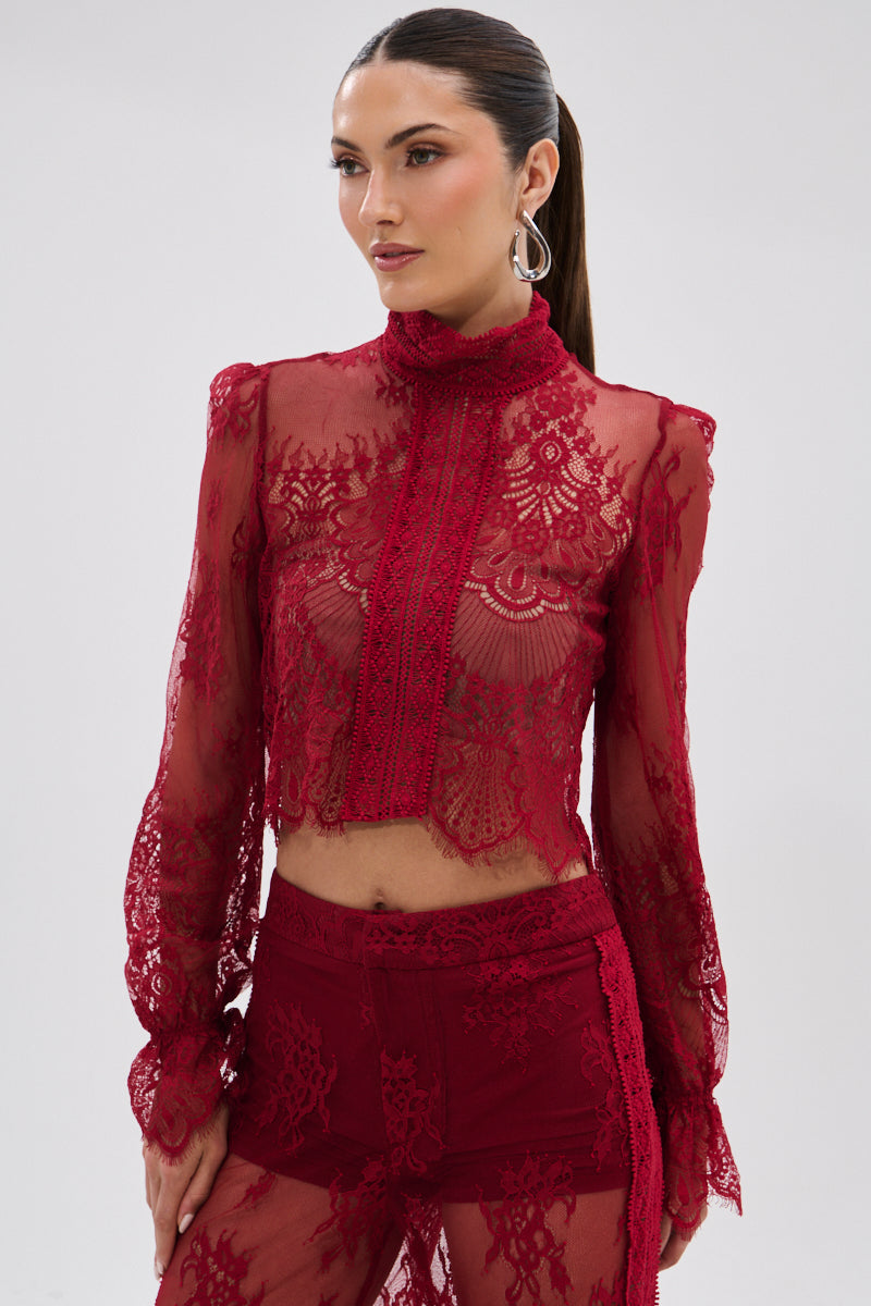 A woman models the LOVE LOST LACE BLOUSE, a sheer red floral lace, long-sleeve top paired with matching high-waisted shorts. Her sleek ponytail and hoop earrings lend a subtle gothic touch against a plain background.