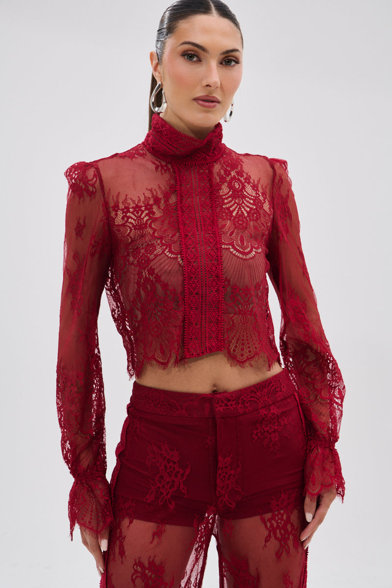 A woman wears the LOVE LOST LACE BLOUSE, a sheer, floral lace, long-sleeve red top paired with matching high-waisted lace pants. Her pulled-back hair and the semi-transparent outfit create a subtle gothic effect on a plain background.