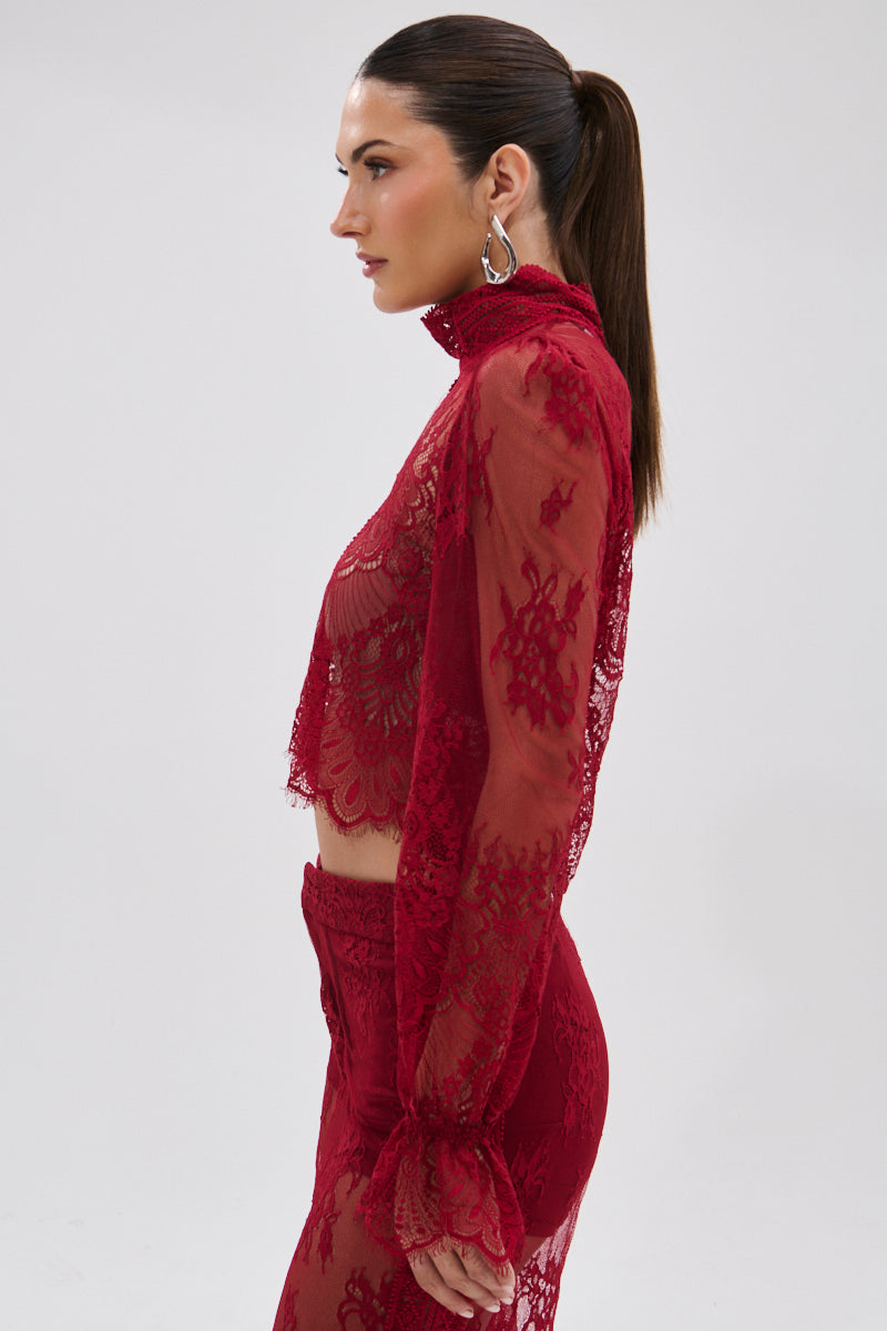 A woman with long brown hair in a ponytail wears the LOVE LOST LACE BLOUSE—featuring sheer floral lace, long sleeves, and a high neck—with matching red lace pants, shown in profile against a plain light background.