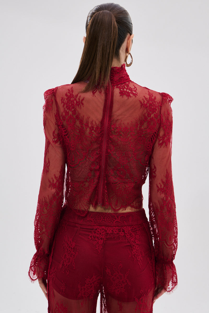 A woman with long brown hair in a ponytail is seen from behind, wearing the sheer floral LOVE LOST LACE BLOUSE, featuring intricate patterns, scalloped edges, and a subtle gothic finish.