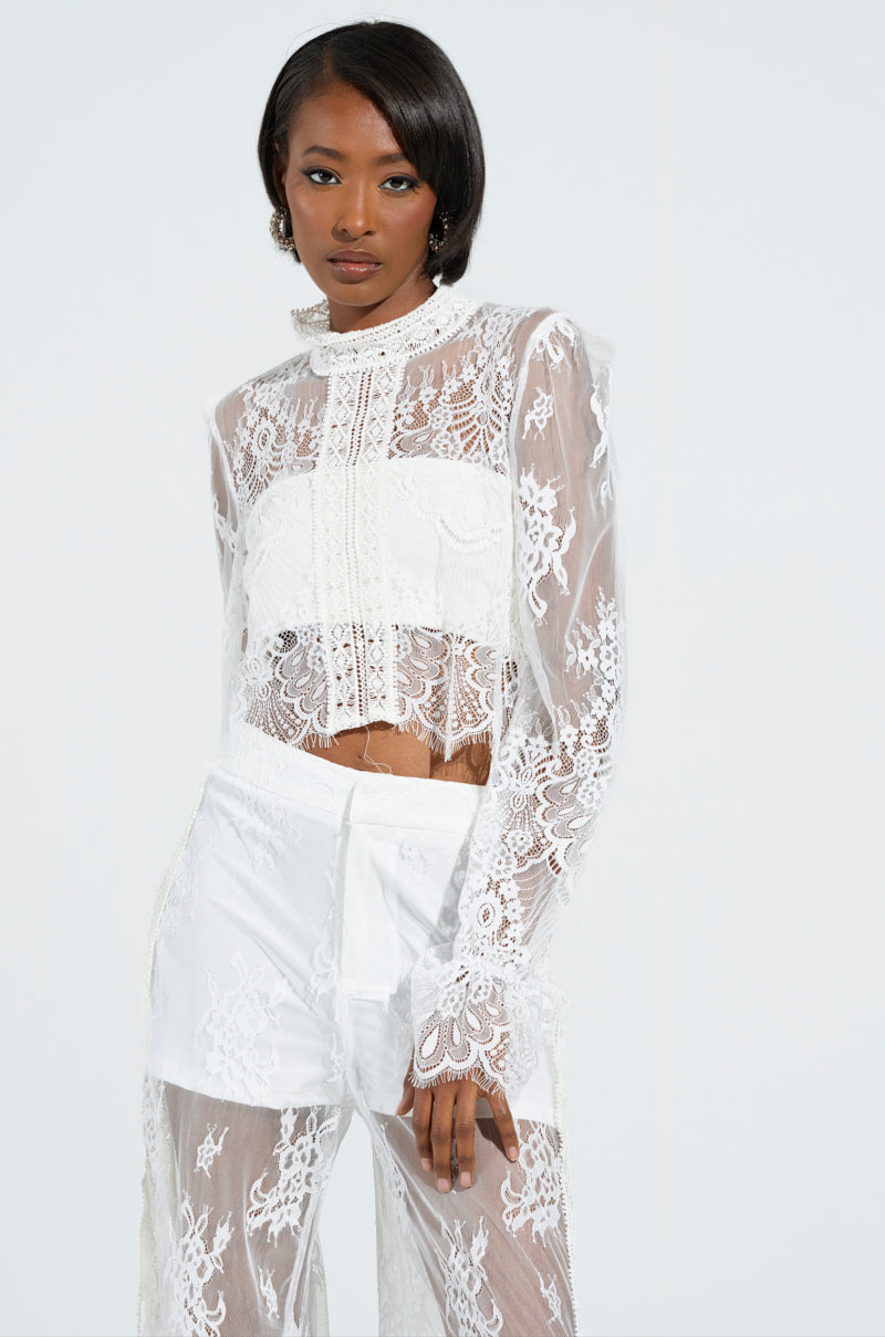 A woman poses in the elegant MISS ME YET LACE BLOUSE, showcasing its sheer white fabric with intricate floral lace patterns. The blouse features long sleeves and a high collar. Her neatly styled dark hair complements the plain light background.