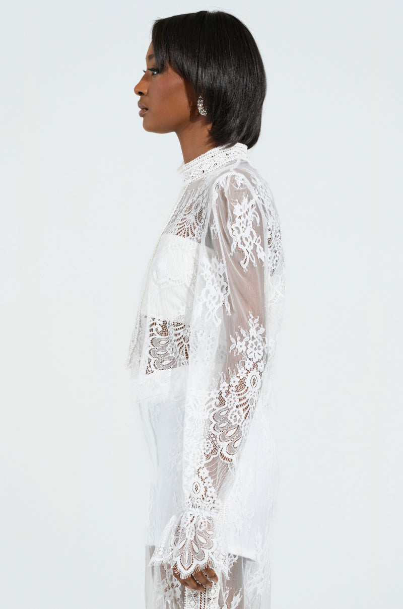 A woman in profile against a pale background wears the MISS ME YET LACE BLOUSE, its intricate floral lace patterns echoing delicacy. Her short, styled hair complements the cropped silhouette as she gazes forward with a calm expression.