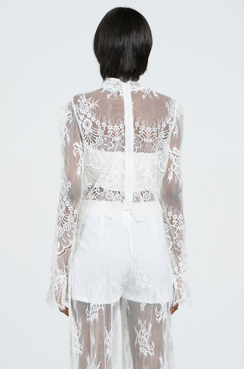 The individual is dressed in the MISS ME YET LACE BLOUSE, a sheer, long-sleeved white lace creation that showcases a cropped silhouette from the back. The blouse is adorned with intricate floral lace patterns and features a high neckline.