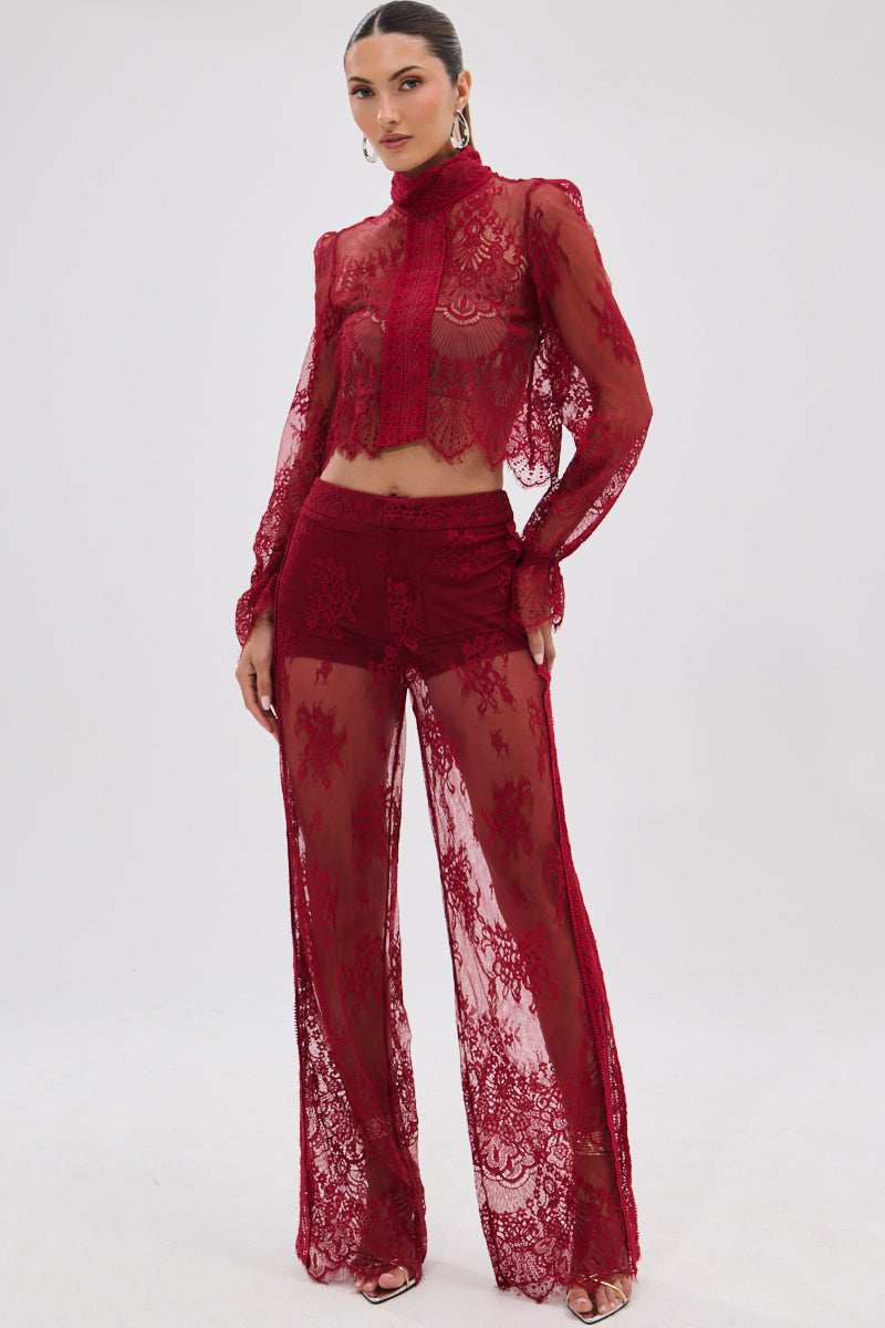 A woman wears the LOVE LOST LACE PANT, a sheer red floral lace two-piece with a high-neck long sleeve crop top and wide-leg, high-rise pants. She accessorizes with hoop earrings, high heels, and has her hair pulled back.