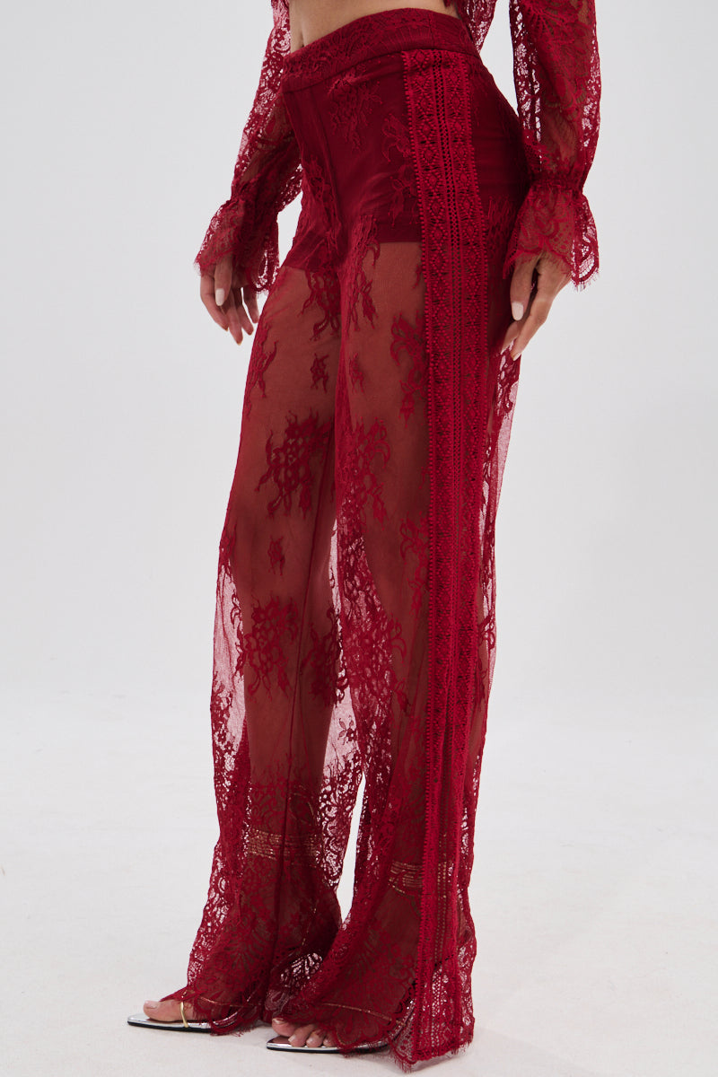 Wearing the LOVE LOST LACE PANT, a sheer, high-rise red lace pant with intricate floral patterns, paired with a matching lace top and open-toe heels that reveal the legs and midriff.