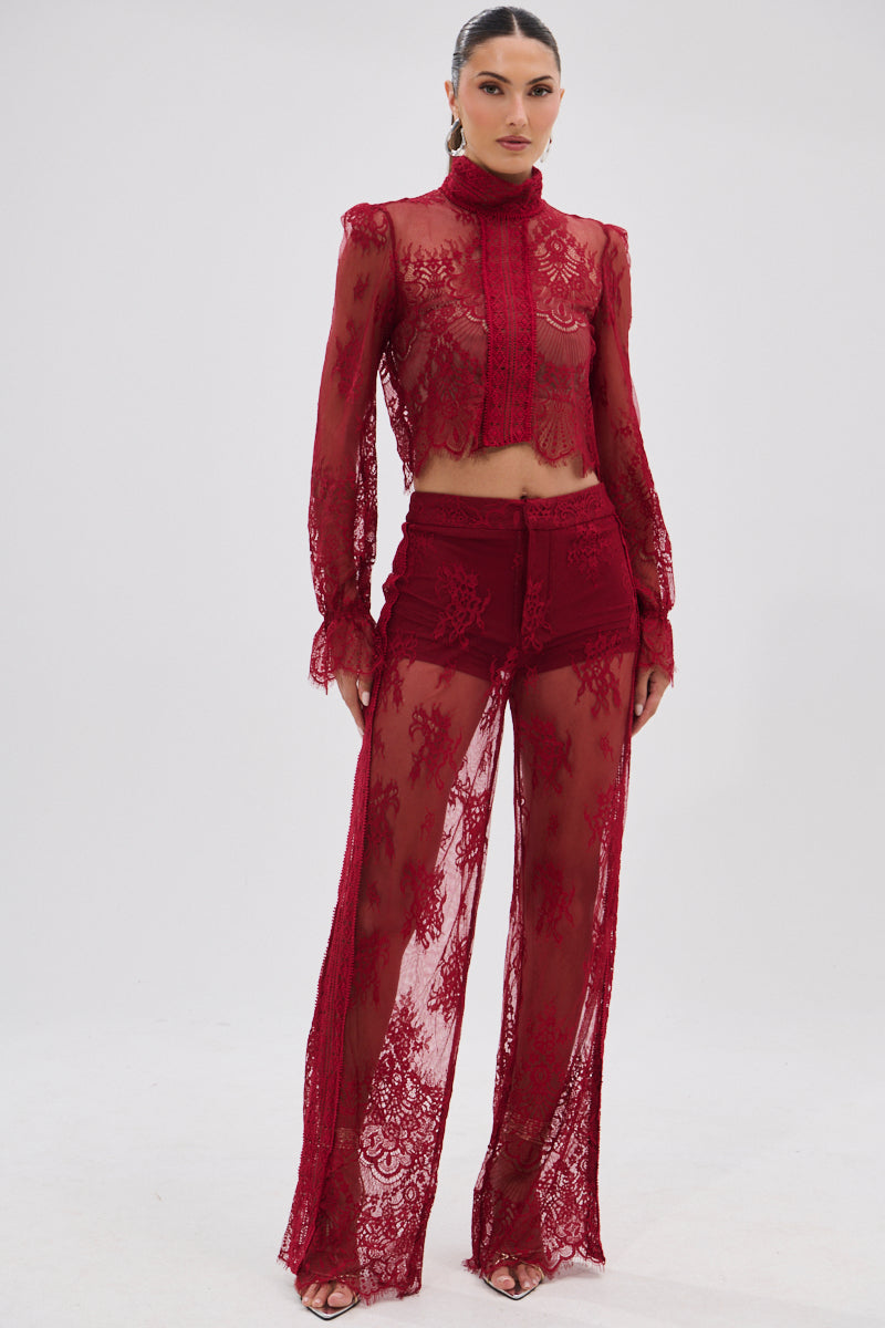 A woman stands confidently against a white background wearing the LOVE LOST LACE PANT, a sheer, dark red floral lace high-rise pant paired with a matching high-neck long-sleeve crop top.