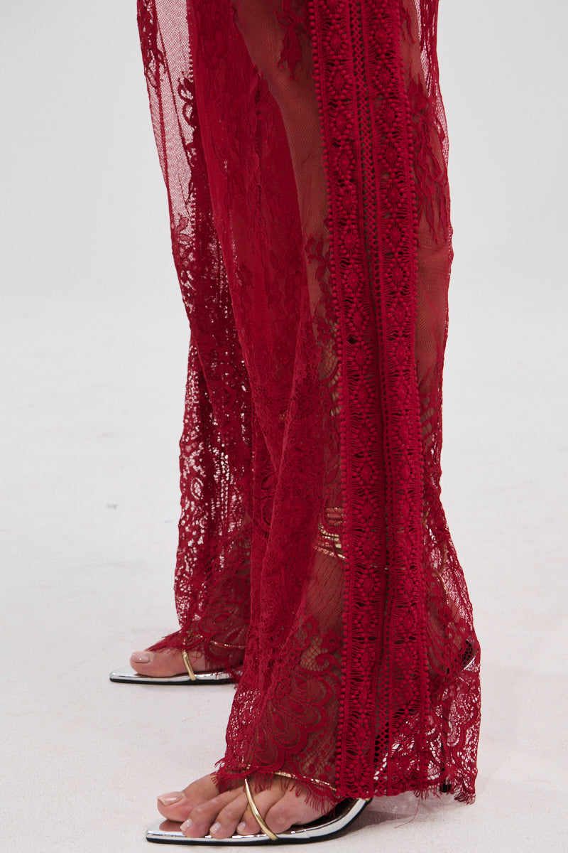 A close-up of a person wearing the LOVE LOST LACE PANT—sheer, floral lace in a high-rise fit—paired with gold-strapped open-toed sandals. Only their feet and lower legs appear against a light background.