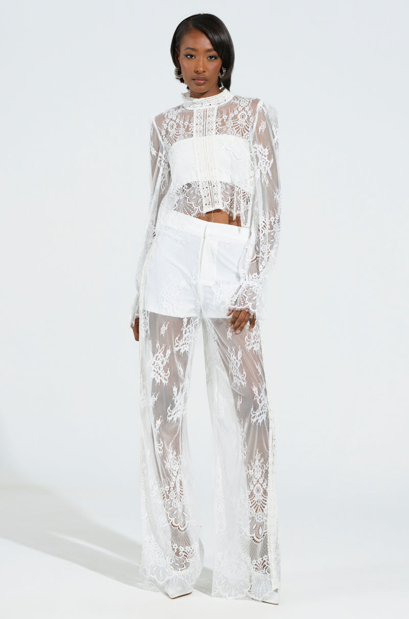 A person is wearing the captivating "MISS ME YET LACE TROUSER" ensemble, featuring a sheer floral lace long-sleeve top paired with high-rise wide-leg trousers. The intricate lace design adds elegance to the outfit, beautifully contrasted against a plain light gray background.