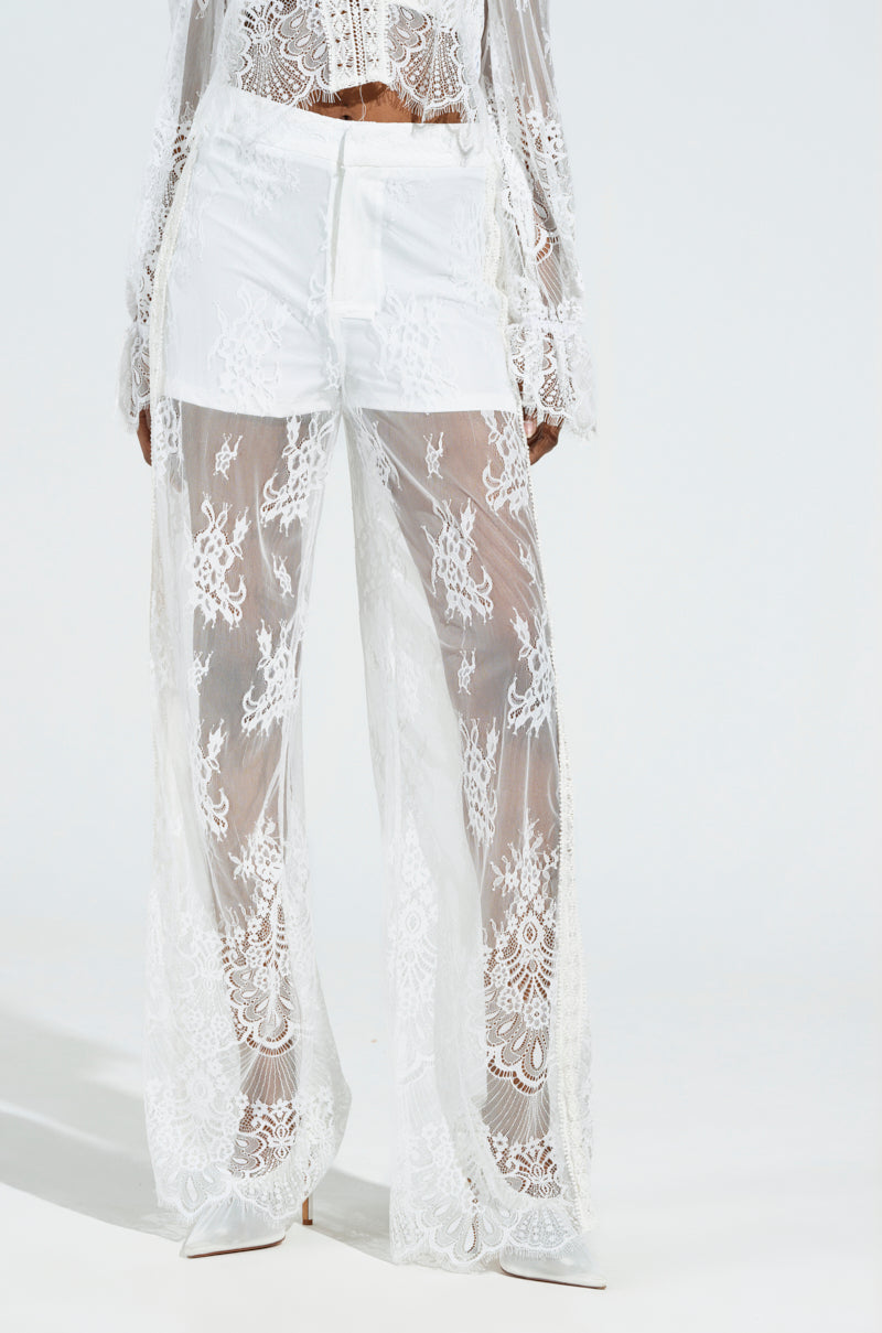 The MISS ME YET LACE TROUSER sits high-rise, featuring intricate floral lace patterns over white shorts, paired with a matching lace top. This elegant ensemble is completed with white shoes against a light background, showcasing the sophistication of the trouser design.