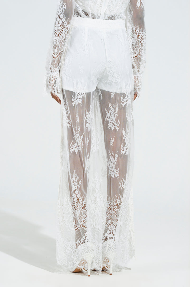 A person wearing the MISS ME YET LACE TROUSER set, featuring intricate sheer white lace with delicate floral patterns, stands with their back to us. The ensemble includes a long-sleeved top and matching high-rise lace trousers, both showcasing exquisite lacework.