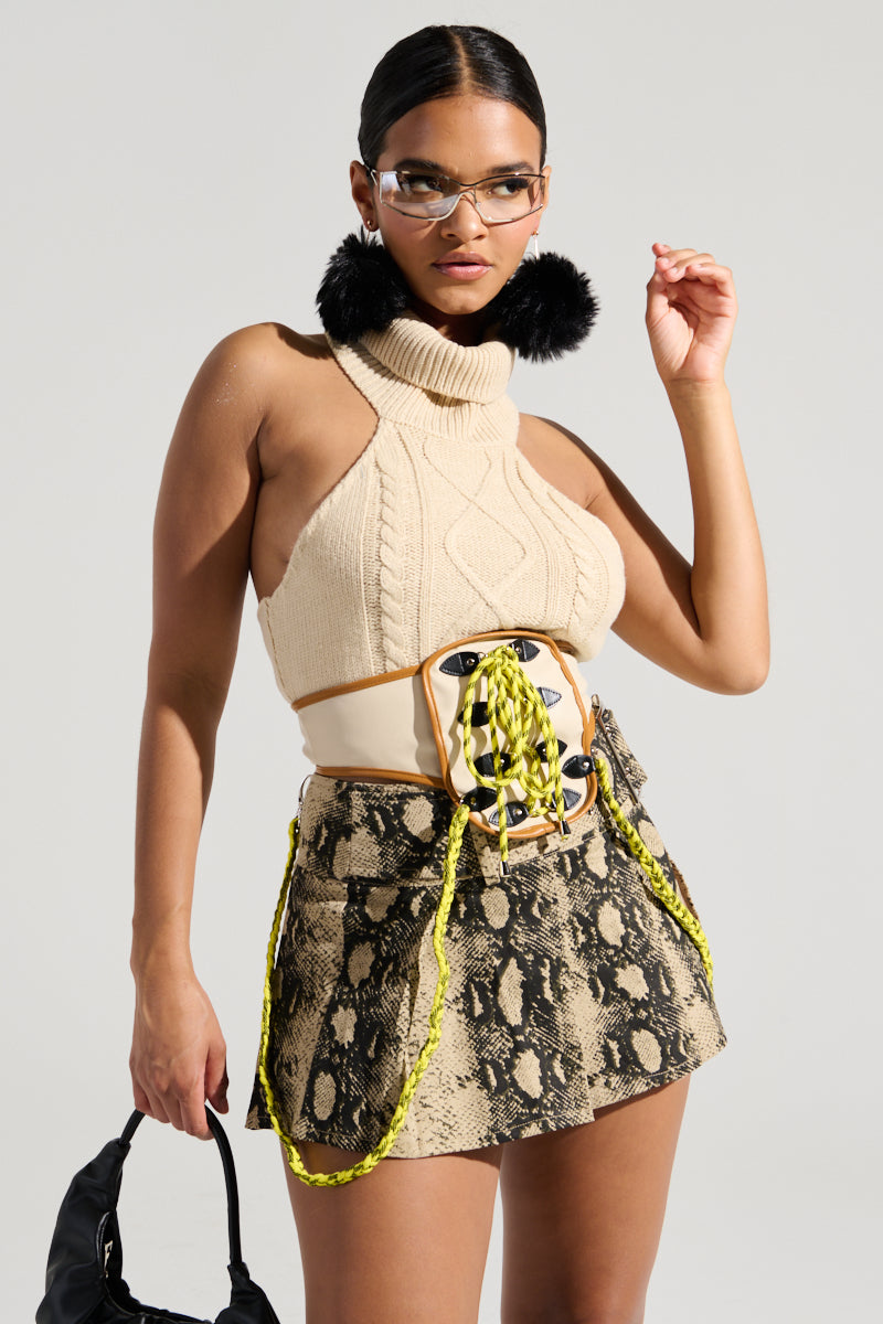 A woman wearing the OUT OF HIS LEAGUE sleeveless fashion top in beige pairs it elegantly with a snakeskin-patterned skirt, complemented by stylish yellow accessories. She accessorizes with black fuzzy earrings, chic glasses, and carries a sleek black handbag. Her confident pose against a neutral background radiates elegance.