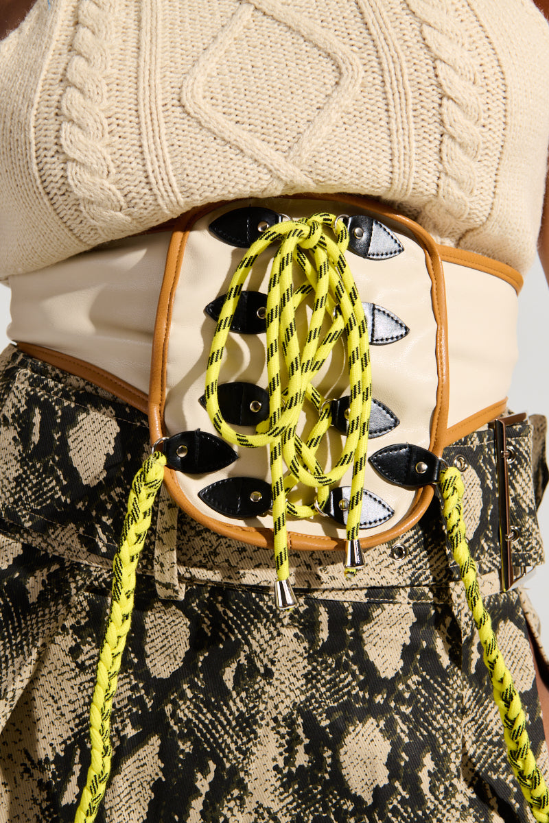 A close-up reveals someone wearing the OUT OF HIS LEAGUE SLEEVELESS FASHION TOP crafted from cream knit fabric, paired with a wide beige faux leather corset belt highlighted by neon yellow laces. The outfit is completed with a snakeskin-patterned skirt, showcasing an eye-catching textured panel design and contrasting colors.