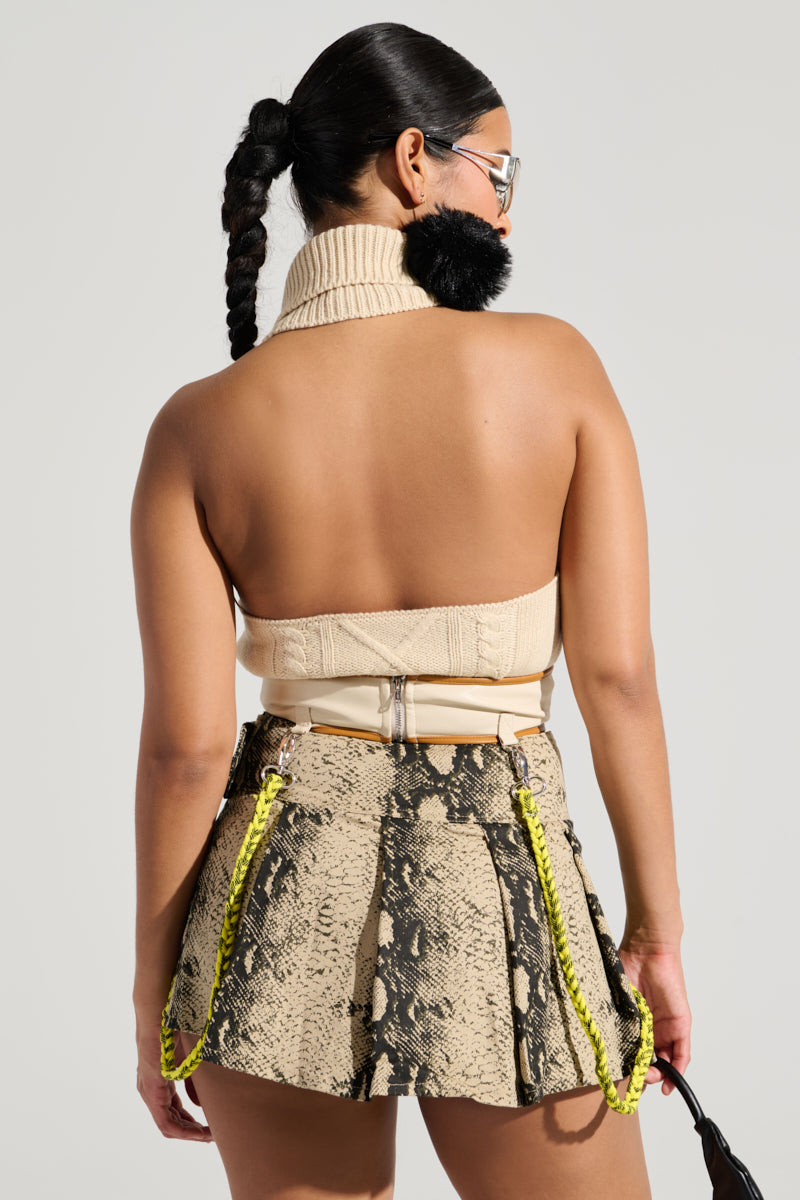 The individual with braided hair is turned away, highlighting their back while wearing the OUT OF HIS LEAGUE SLEEVELESS FASHION TOP in beige. They pair this top with a snakeskin-patterned skirt featuring yellow chain accents, complemented by a textured panel design and a black fur accessory draped around the neck for added chicness.