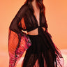 A woman poses against an orange background wearing THE JULIE BLOUSE, a sheer mesh top with floral-patterned sleeves and hem, a deep V-neck, waist ties, and wide flowing sleeves for a dramatic look.