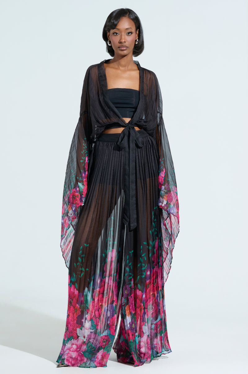 A person dons a sheer black ensemble with wide sleeves, highlighting THE JULIE BLOUSE's vibrant floral pattern below. The outfit includes a tie-front cropped blouse over a black bandeau and matching flowing pants, all set against a clean white background that enhances the colorful design.