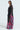 A model poses against a light background in THE JULIE BLOUSE, a sheer, black floral piece adorned with vibrant pink and purple flowers. The blouse features long, flowing sleeves and a tie-front design, creating an elegant, layered appearance reminiscent of a gown.