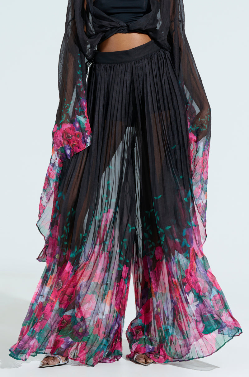 Dressed in THE JULIE TROUSER with flowy, wide-leg chiffon pants featuring a black base and vibrant pink floral pattern, paired with a sheer top adorned with long, draping sleeves, this outfit radiates a stylish and elegant look.