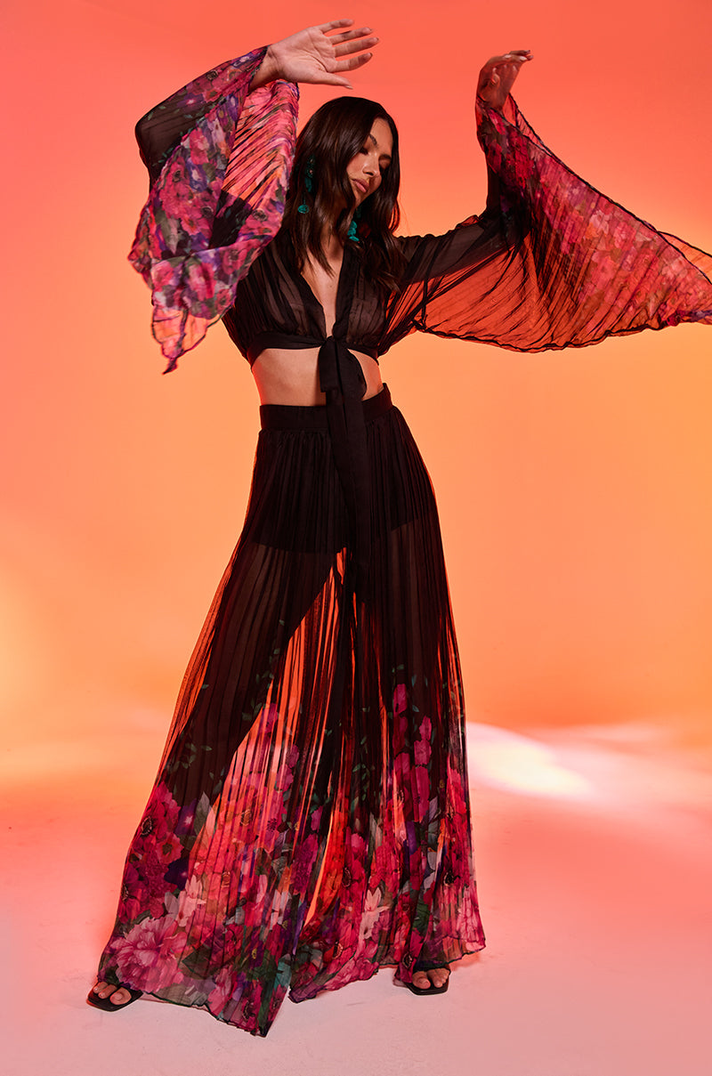 A woman poses against an orange backdrop in THE JULIE TROUSER—sheer black wide leg pants with vivid pink and purple florals. The high rise mesh and flowing silhouette add elegance as she raises her arms gracefully and turns her head.