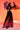 A woman poses against an orange backdrop in THE JULIE TROUSER—sheer black wide leg pants with vivid pink and purple florals. The high rise mesh and flowing silhouette add elegance as she raises her arms gracefully and turns her head.