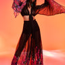 A woman poses against an orange backdrop in THE JULIE TROUSER—sheer black wide leg pants with vivid pink and purple florals. The high rise mesh and flowing silhouette add elegance as she raises her arms gracefully and turns her head.
