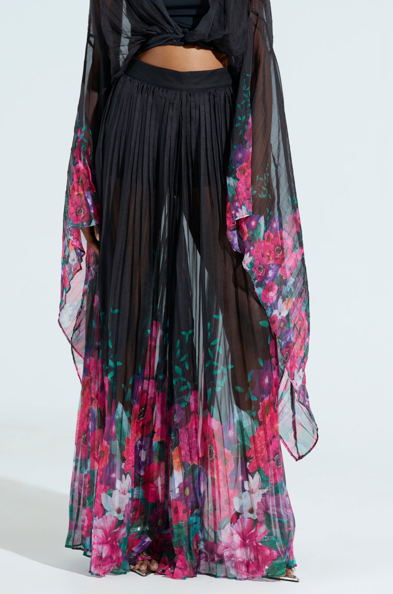 Someone is wearing a sheer black outfit with THE JULIE TROUSER, featuring vibrant floral print pants in shades of pink, red, and green, with a wide-leg silhouette and pleated fabric. The background is a plain, light color.