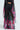 Someone is wearing a sheer black outfit with THE JULIE TROUSER, featuring vibrant floral print pants in shades of pink, red, and green, with a wide-leg silhouette and pleated fabric. The background is a plain, light color.