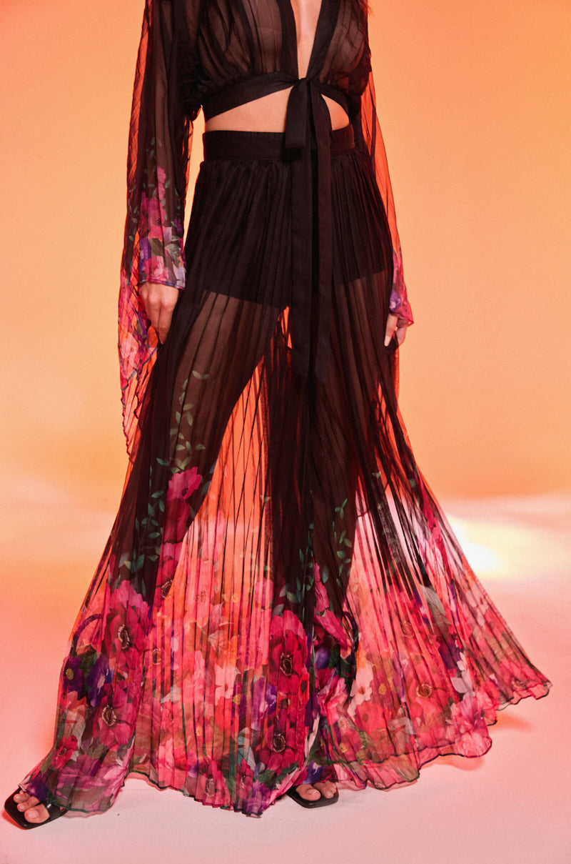 A person stands against an orange background wearing THE JULIE TROUSER—high-rise, wide-leg pants with vibrant pink floral prints—paired with a sheer black top and black sandals.