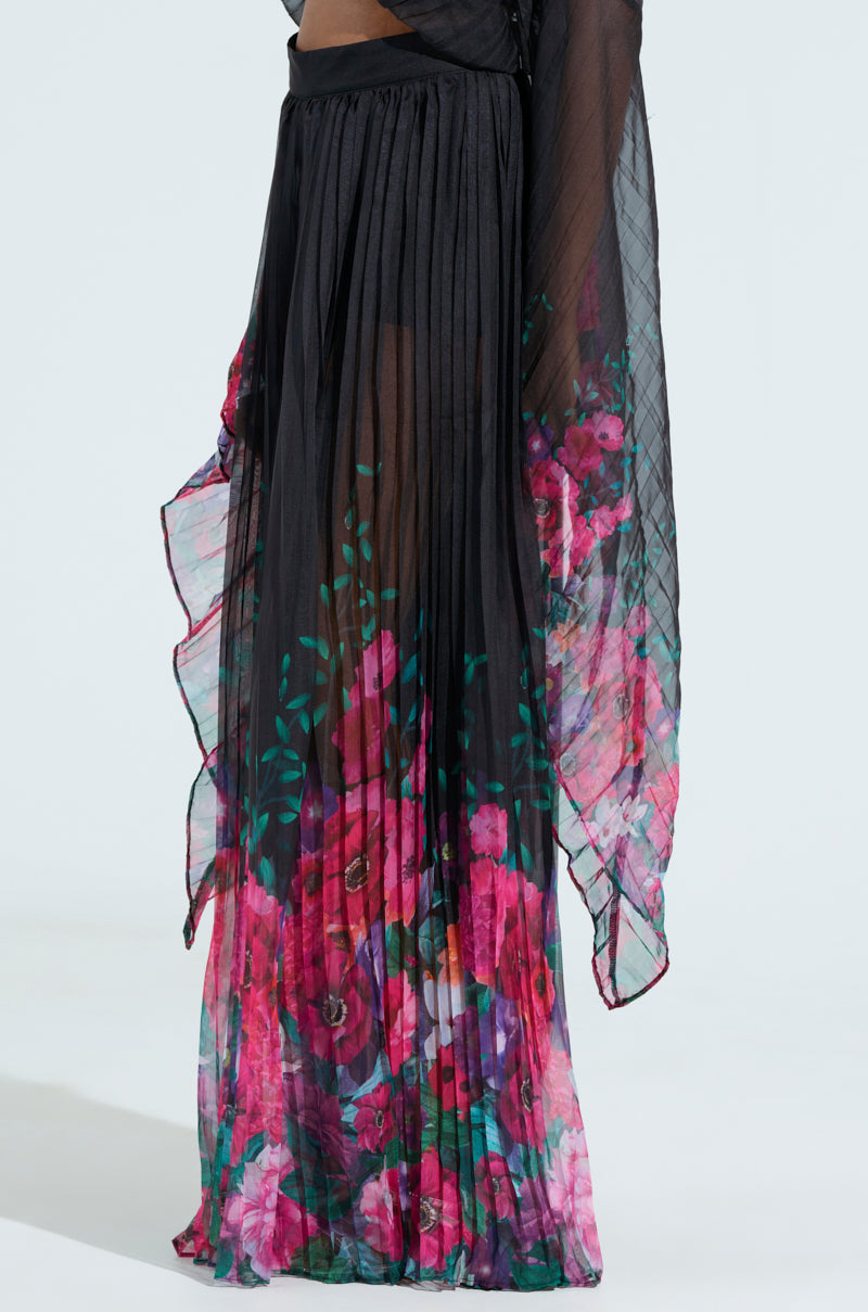 A sheer black pleated dress named THE JULIE TROUSER, featuring a high-rise design and vibrant pink and green floral patterns. This semi-transparent garment flows elegantly, creating a soft silhouette against the light background.