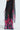 A sheer black pleated dress named THE JULIE TROUSER, featuring a high-rise design and vibrant pink and green floral patterns. This semi-transparent garment flows elegantly, creating a soft silhouette against the light background.