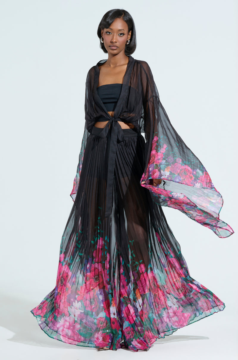 A woman confidently stands against a plain white background wearing THE JULIE TROUSER, featuring vibrant pink floral print and a wide-leg silhouette. She pairs it with a tied top, opting for minimal accessories to complement the flowing, sheer black outfit.