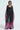 A person in THE JULIE TROUSER, a high-rise design with vibrant pink and green floral patterns, paired with a flowing black outfit featuring a sheer overlay, wide sleeves, and a knotted front. They stand against a plain background.