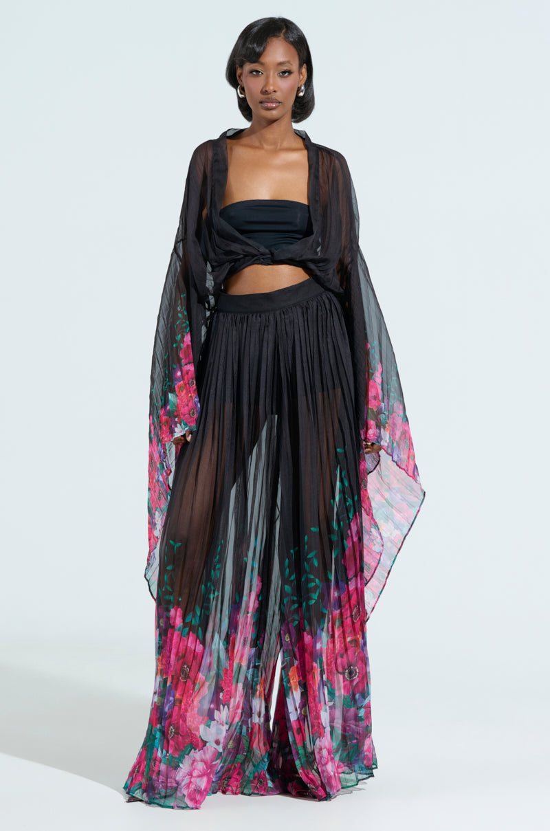 A woman wears THE JULIE TROUSER, a flowy, sheer black ensemble with vibrant floral print. The outfit includes a bandeau top and high-rise wide-leg silhouette, her straight hair complementing the look against a plain white background.