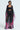 A woman wears THE JULIE TROUSER, a flowy, sheer black ensemble with vibrant floral print. The outfit includes a bandeau top and high-rise wide-leg silhouette, her straight hair complementing the look against a plain white background.