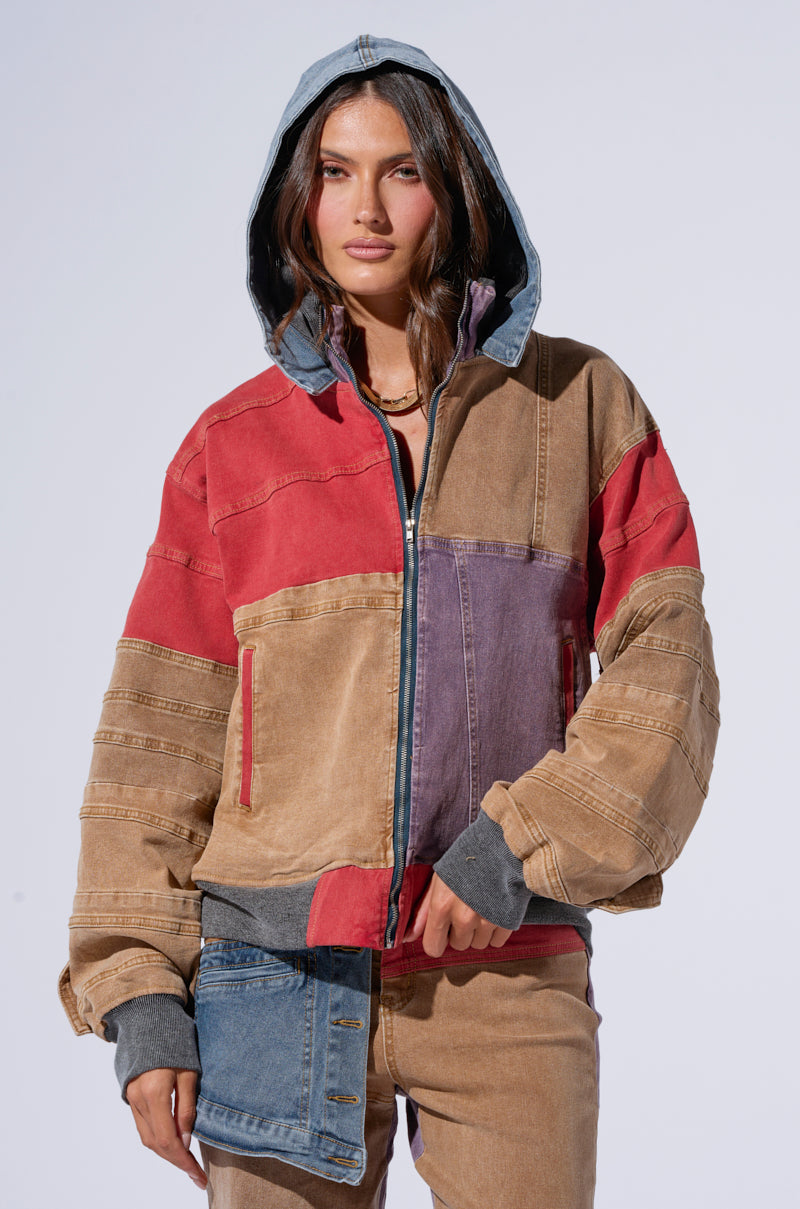 A person stands against a plain background wearing the SUPERNATURAL PATCHWORK DENIM BOMBER from AKIRA Label, featuring various shades of brown, red, and denim blue. The jacket includes a hood and several zippers. The person has long brown hair and a neutral expression.