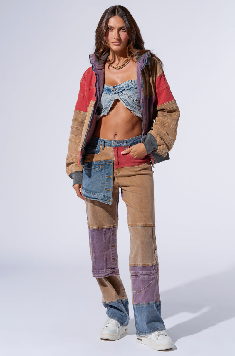 A woman stands against a plain background wearing a colorful patchwork ensemble. She has on an AKIRA Label multicolored jacket with red, purple, and brown patches, paired with a blue patterned crop top. Her outfit features the Supernatural Patchwork Jeans, combining purple, brown, and blue denim patches. To complete her look, she sports white sneakers.
