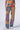 A person is modeling the Supernatural Patchwork Jeans from the AKIRA Label, which feature an eclectic mix of multi-colored denim patches in shades of blue, brown, purple, and red for a vintage-inspired look. The high-rise waist perfectly complements their white sneakers and cropped fringed top. The plain, light background keeps the focus on their stylish ensemble.