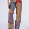 A person is modeling the Supernatural Patchwork Jeans from the AKIRA Label, which feature an eclectic mix of multi-colored denim patches in shades of blue, brown, purple, and red for a vintage-inspired look. The high-rise waist perfectly complements their white sneakers and cropped fringed top. The plain, light background keeps the focus on their stylish ensemble.