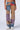 A person wearing SUPERNATURAL PATCHWORK JEANS, which showcase an array of colors like blue, purple, red, and brown patches. The high-rise waist adds a stylish touch to their outfit. Paired with white sneakers against a plain white background.