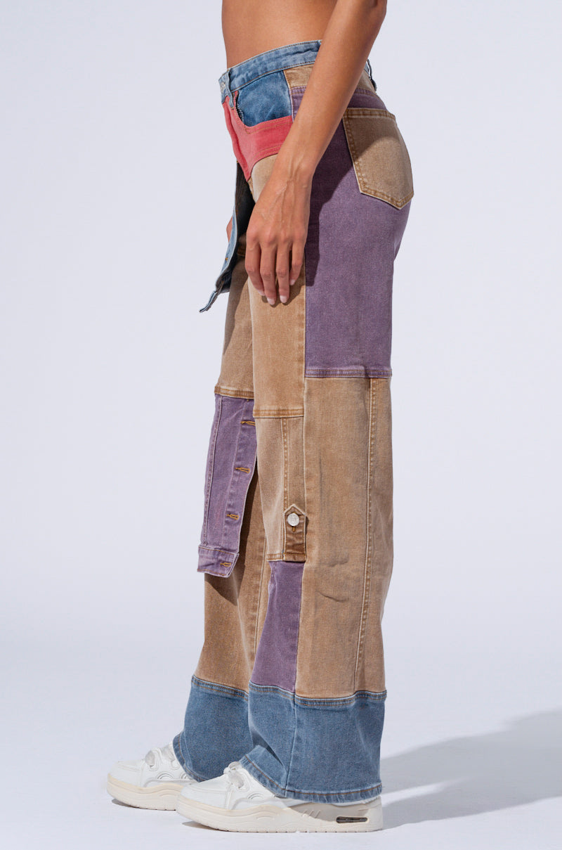 A person wearing AKIRA Label SUPERNATURAL PATCHWORK JEANS featuring a high-rise waist in various shades of brown, blue, red, and purple stands against a plain light gray background. The jeans showcase a mix of fabrics and colors, complemented by white sneakers.