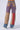 A person wearing the SUPERNATURAL PATCHWORK JEANS from AKIRA Label, featuring a high-rise waist, stands facing away. The color-blocked wide-leg pants exhibit a blend of purple, tan, red, and blue shades with prominent patchwork seams and two back pockets. The look is finished off with white sneakers against a plain white backdrop.